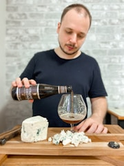 A man pours a dark beer from a bottle into a glass on a wooden table. Next to the glass is a block of blue cheese and several pieces of the cheese are crumbled nearby. The background features a textured gray brick wall.
