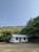 A small, simple house with a white and grey exterior is situated in a rural setting. Surrounding the house are lush green trees and hills in the background under a clear blue sky. The ground appears dry and grassy.