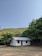 A small, simple house with a white and grey exterior is situated in a rural setting. Surrounding the house are lush green trees and hills in the background under a clear blue sky. The ground appears dry and grassy.