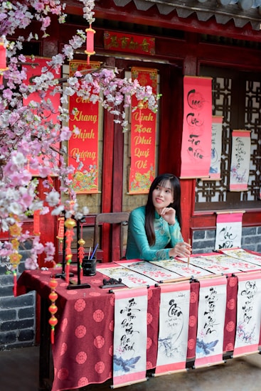 A woman in a traditional teal dress is sitting at a table decorated with red cloth and calligraphy. The table is set in front of a building with red and gold decorative items, including hanging scrolls with Chinese characters and cherry blossom branches. The setting has a festive and cultural atmosphere.
