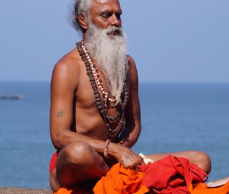 topless man sitting on brown sand near body of water during daytime