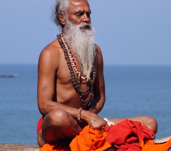 topless man sitting on brown sand near body of water during daytime