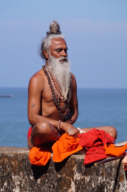 topless man sitting on brown sand near body of water during daytime