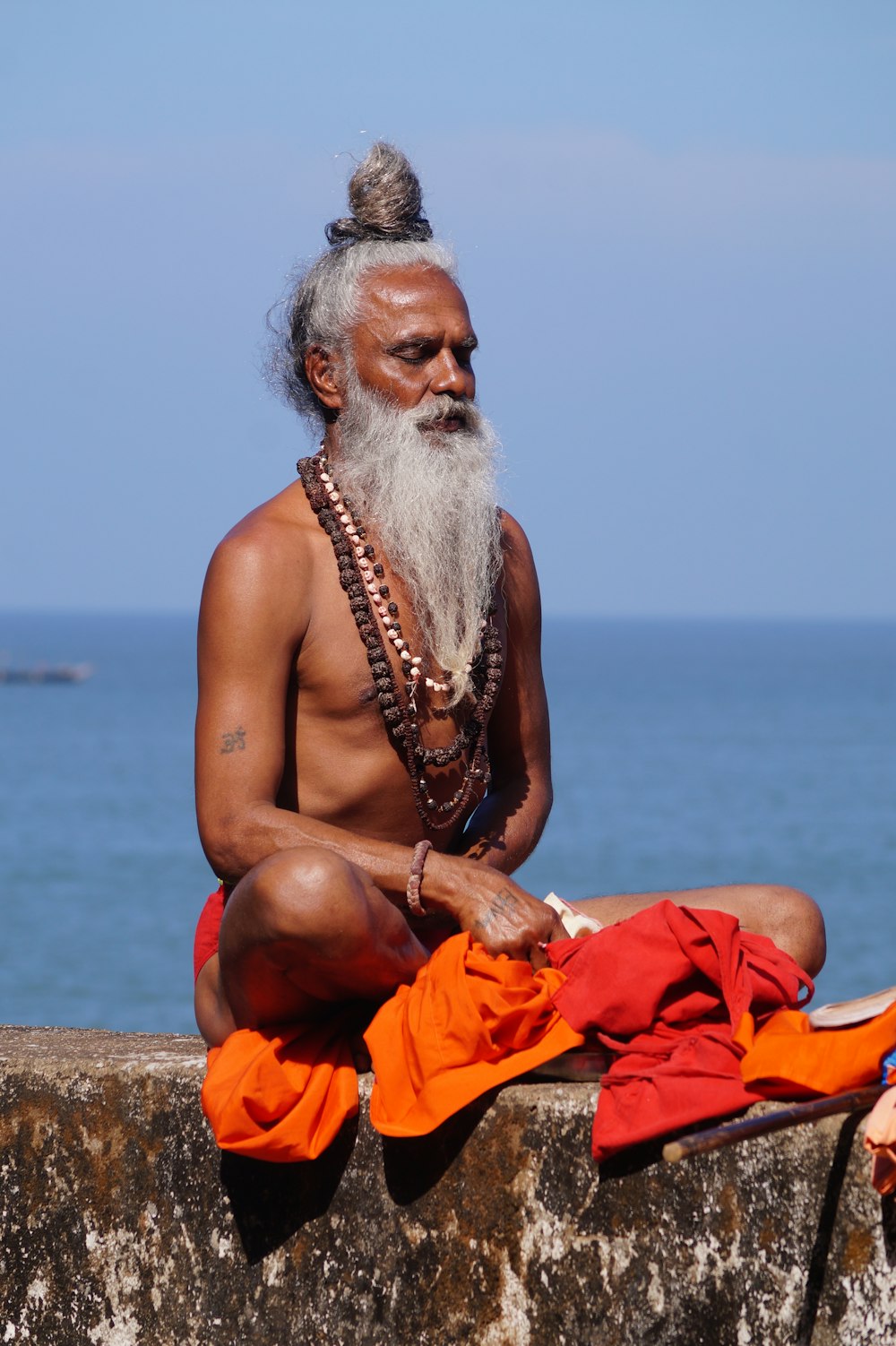 topless man sitting on brown sand near body of water during daytime