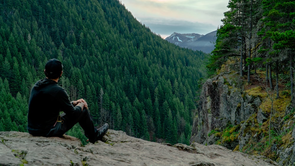 man in black jacket and black pants sitting on rock near green trees during daytime