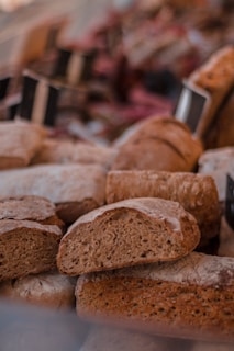 Various types of freshly baked bread are displayed, including loaves and sliced pieces with a rustic and hearty appearance. The backdrop suggests a market or bakery setting, with blurred signs in the background.