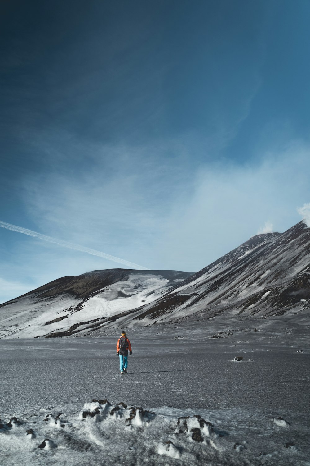 파란색 재킷과 파란색 데님 청바지를 입은 사람이 회색 산 근처 회색 모래 위에 서 있습니다.