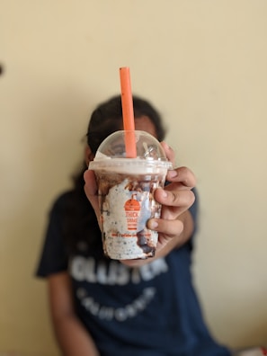 A person holds a creamy chocolate milkshake in a transparent plastic cup with a domed lid. The cup features branding from 'The Thick Shake Factory' and contains visible chocolate syrup swirls and cookie pieces. An orange straw protrudes from the lid.