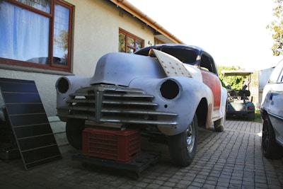 A vintage car in the process of restoration is positioned in a driveway next to a house. The car's body is partially sanded and prepared for further repair, with the front lacking headlights and covered in primer. The front of the car is lifted and supported by a red plastic crate. Next to the car on the left, there is a solar panel leaning against the house wall, and another vehicle is visible in the background.