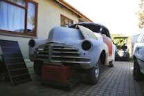 A vintage car in the process of restoration is positioned in a driveway next to a house. The car's body is partially sanded and prepared for further repair, with the front lacking headlights and covered in primer. The front of the car is lifted and supported by a red plastic crate. Next to the car on the left, there is a solar panel leaning against the house wall, and another vehicle is visible in the background.