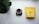 A white cup filled with coffee beans sits on a digital scale displaying zero grams. To the right, there is a yellow coffee bag with branding text, placed on a mostly white surface. A large green plant leaf is partially visible in the top left corner.