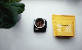 A white cup filled with coffee beans sits on a digital scale displaying zero grams. To the right, there is a yellow coffee bag with branding text, placed on a mostly white surface. A large green plant leaf is partially visible in the top left corner.