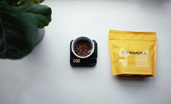 A white cup filled with coffee beans sits on a digital scale displaying zero grams. To the right, there is a yellow coffee bag with branding text, placed on a mostly white surface. A large green plant leaf is partially visible in the top left corner.