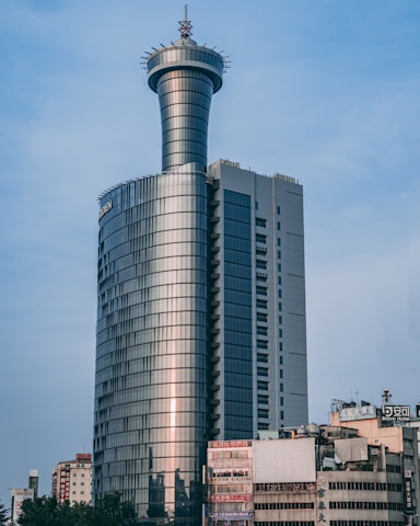 A tall, modern skyscraper with a unique cylindrical tower on top. The building has a sleek, reflective glass facade and a vertical structure. Surrounding it are several older, shorter commercial buildings with visible signs and advertisements.