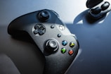 A black gaming controller with colorful buttons labeled X, Y, A, and B is positioned on a flat surface next to wireless earbuds in their charging case. Sunlight casts shadows, emphasizing the controller's sleek design.