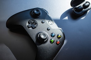 A black gaming controller with colorful buttons labeled X, Y, A, and B is positioned on a flat surface next to wireless earbuds in their charging case. Sunlight casts shadows, emphasizing the controller's sleek design.