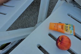 A tube of sunscreen and a ripe peach are positioned on a white lounge chair, with parts of a person's leg and shoe visible at the edge.