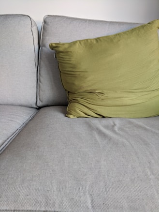 A close-up view of a light gray fabric sofa with a green pillow resting on it. The sofa cushions appear comfortable and slightly textured, while the pillow is soft and slightly wrinkled, positioned at an angle.