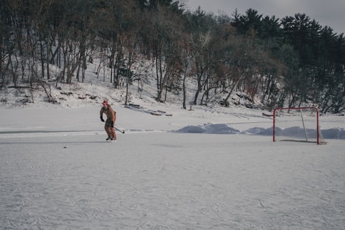 A person is ice skating and playing hockey on a snow-covered outdoor rink surrounded by trees. A hockey goal stands to the right of the skater. The background consists of a snowy wooded area with leafless and evergreen trees.