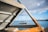 A small sailboat is floating on a calm sea, framed by a triangular opening created by a rusted metal structure in the foreground. The sky is partly cloudy with patches of blue.