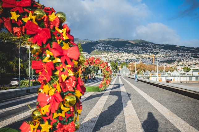 A street adorned with colorful holiday decorations, featuring large festive arches made of red bows, golden stars, and green leaves. The road stretches into the distance with mountainous scenery and a partly cloudy sky in the background.