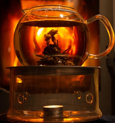 A transparent glass teapot containing a blooming flower tea sits atop a glass base. The warm glow from a single tea light candle beneath creates a cozy atmosphere. In the background, hints of a fireplace or a warm light source add a soothing orange and yellow glow.