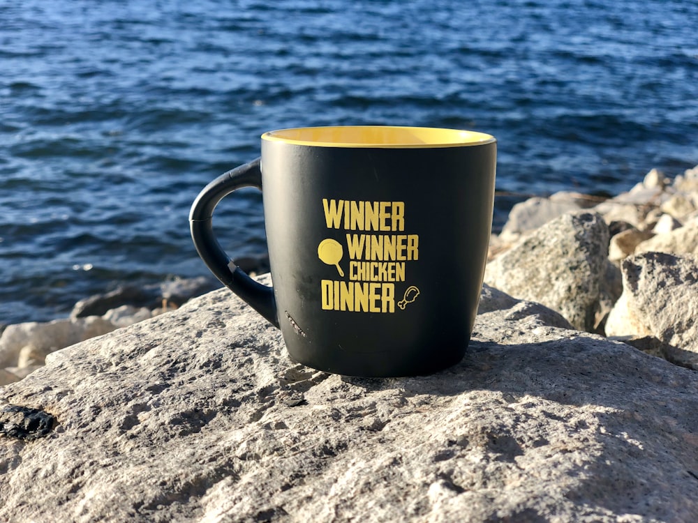 black and yellow ceramic mug on gray rock near body of water during daytime