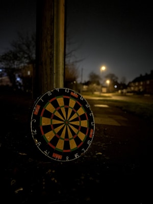 A dartboard leans against a post on a dimly lit street at night. The scene is quiet with soft streetlights illuminating the distant houses and trees lining the sidewalk. The dartboard is colorful, with red, green, and black sections.