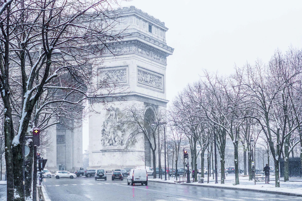 The Arc de Triomphe in falling snow, framed by bare snow-covered trees along the avenue, with a few cars on the wet road