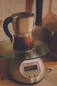 A metallic stovetop coffee maker sits on a digital kitchen scale, with a backdrop of warm-toned kitchen utensils and ceramic tiles. The scale displays a reading, and both the coffee maker and the scale appear to be well-used.