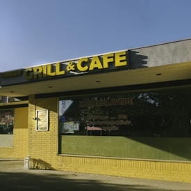 A storefront with a yellow brick exterior and a large sign reading 'GRILL & CAFE'. The sign features bold yellow letters mounted on a gray background. Below the sign, there are large windows with menus and advertisements displayed. The sunlight casts shadows on the ground and partially on the building.
