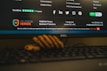 A computer monitor displays a webpage from HelpDesk Heroes, featuring a contact address and email. Social media icons including Facebook, LinkedIn, Twitter, Instagram, and Pinterest are visible. The screen also shows a Trustpilot rating. A stack of cookies is partially visible at the bottom of the image, and part of a keyboard can be seen in the foreground.