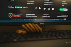 A computer monitor displays a webpage from HelpDesk Heroes, featuring a contact address and email. Social media icons including Facebook, LinkedIn, Twitter, Instagram, and Pinterest are visible. The screen also shows a Trustpilot rating. A stack of cookies is partially visible at the bottom of the image, and part of a keyboard can be seen in the foreground.