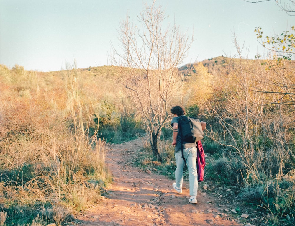 woman in black jacket and blue denim jeans walking on dirt road between brown grass field