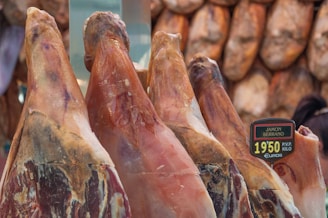 Several cured ham legs, known as jamon serrano, are displayed upright in a shop setting. A price tag is visible, indicating a price per kilogram. The background features hanging cured meats, contributing to an overall rustic and traditional market atmosphere.