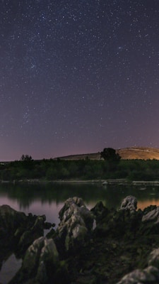 A calm, starlit night sky reflects over a tranquil body of water, surrounded by rocky formations. The distant horizon features gentle hills and sparse trees, bathed in soft, ambient light.