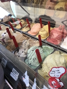 A display of assorted ice cream flavors in a gelato shop, featuring various colors and textures. Each flavor is in a separate container with red handles sticking out, and there are small signs indicating the names of some flavors. The ice cream is creatively decorated, and a sign advertises homemade special cones.