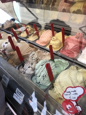 A display of assorted ice cream flavors in a gelato shop, featuring various colors and textures. Each flavor is in a separate container with red handles sticking out, and there are small signs indicating the names of some flavors. The ice cream is creatively decorated, and a sign advertises homemade special cones.