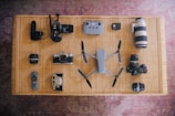 A variety of electronic devices and camera equipment neatly arranged on a woven surface. Visible items include several cameras, camera lenses, a drone with propellers extended, a small remote control, and an audio recording device. The arrangement is symmetrical and organized, showcasing the collection.