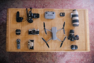 A variety of electronic devices and camera equipment neatly arranged on a woven surface. Visible items include several cameras, camera lenses, a drone with propellers extended, a small remote control, and an audio recording device. The arrangement is symmetrical and organized, showcasing the collection.