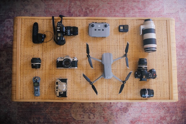 A variety of electronic devices and camera equipment neatly arranged on a woven surface. Visible items include several cameras, camera lenses, a drone with propellers extended, a small remote control, and an audio recording device. The arrangement is symmetrical and organized, showcasing the collection.