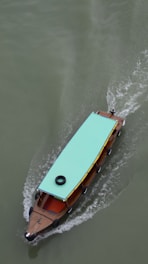 A wooden boat with a turquoise canopy is moving through greenish water, leaving a trail of ripples behind. There is a black tire attached to the top of the canopy.