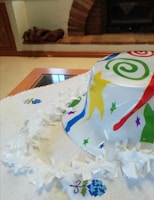 A colorful plastic party hat with swirls and star patterns is placed on a table with a white tablecloth featuring small embroidered blue flowers. The background shows a brick fireplace with stacked wood.