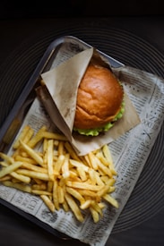 A fresh hamburger with a golden brown bun partially wrapped in paper, accompanied by a generous serving of crispy, golden fries. The meal is presented on a tray lined with paper that resembles newspaper.