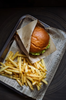 A fresh hamburger with a golden brown bun partially wrapped in paper, accompanied by a generous serving of crispy, golden fries. The meal is presented on a tray lined with paper that resembles newspaper.