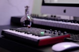 A music studio setup featuring a digital keyboard with a red base is in the foreground. A computer is displayed at the back with an Apple logo on the monitor. A small, reflective bottle sits nearby, adding a metallic sheen. The overall setup suggests a combination of music and technology.