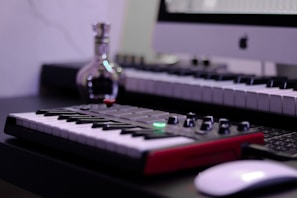 A music studio setup featuring a digital keyboard with a red base is in the foreground. A computer is displayed at the back with an Apple logo on the monitor. A small, reflective bottle sits nearby, adding a metallic sheen. The overall setup suggests a combination of music and technology.