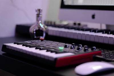 A music studio setup featuring a digital keyboard with a red base is in the foreground. A computer is displayed at the back with an Apple logo on the monitor. A small, reflective bottle sits nearby, adding a metallic sheen. The overall setup suggests a combination of music and technology.