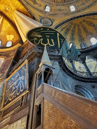 An intricate architectural interior featuring a large circular plaque with Arabic script, ornate gold and blue decorative patterns on the walls and ceiling, and detailed carvings on wooden structures. The light filters through small windows, highlighting the rich colors and textures.