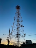 A tall telecommunications tower silhouetted against a clear blue sky during sunset, with various antennas attached at different heights. Power lines and utility poles are visible in the foreground, along with some buildings and trees at the base of the tower.